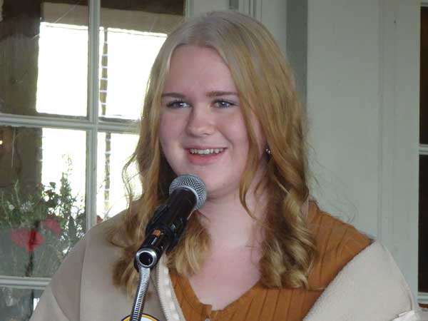 A person with blonde hair speaking into a microphone

Description automatically generated