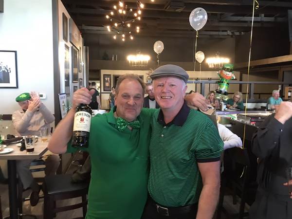 Two men in green shirts holding a bottle of wine

Description automatically generated