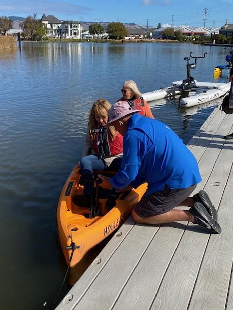 A person kneeling on a dock next to a person in a kayak

Description automatically generated